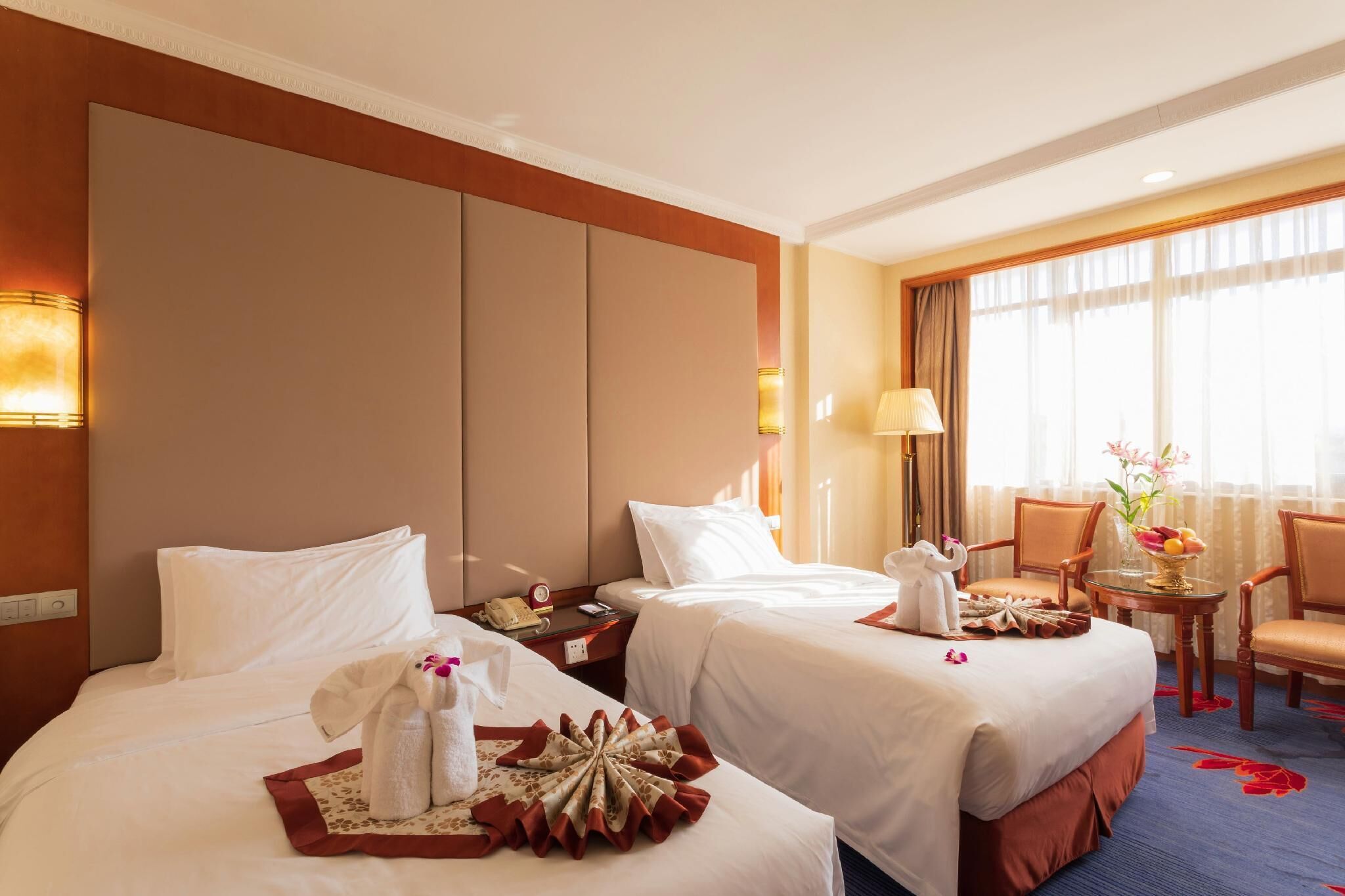Quarto de hotel com duas camas, uma escrivaninha e uma cadeira. Há uma luminária, um vaso com flores e uma fruteira na escrivaninha.