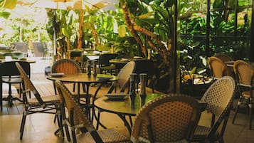 An indoor dining area with wicker chairs and tables, surrounded by large windows and lush greenery.