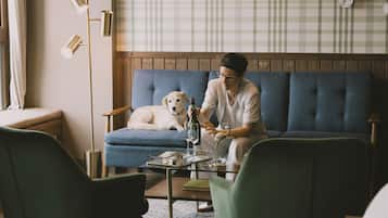 Una sala de estar acogedora con un sofá azul, un sillón verde y una mesa de centro de madera. Un perro está descansando en el sofá, y una persona sentada a la mesa.