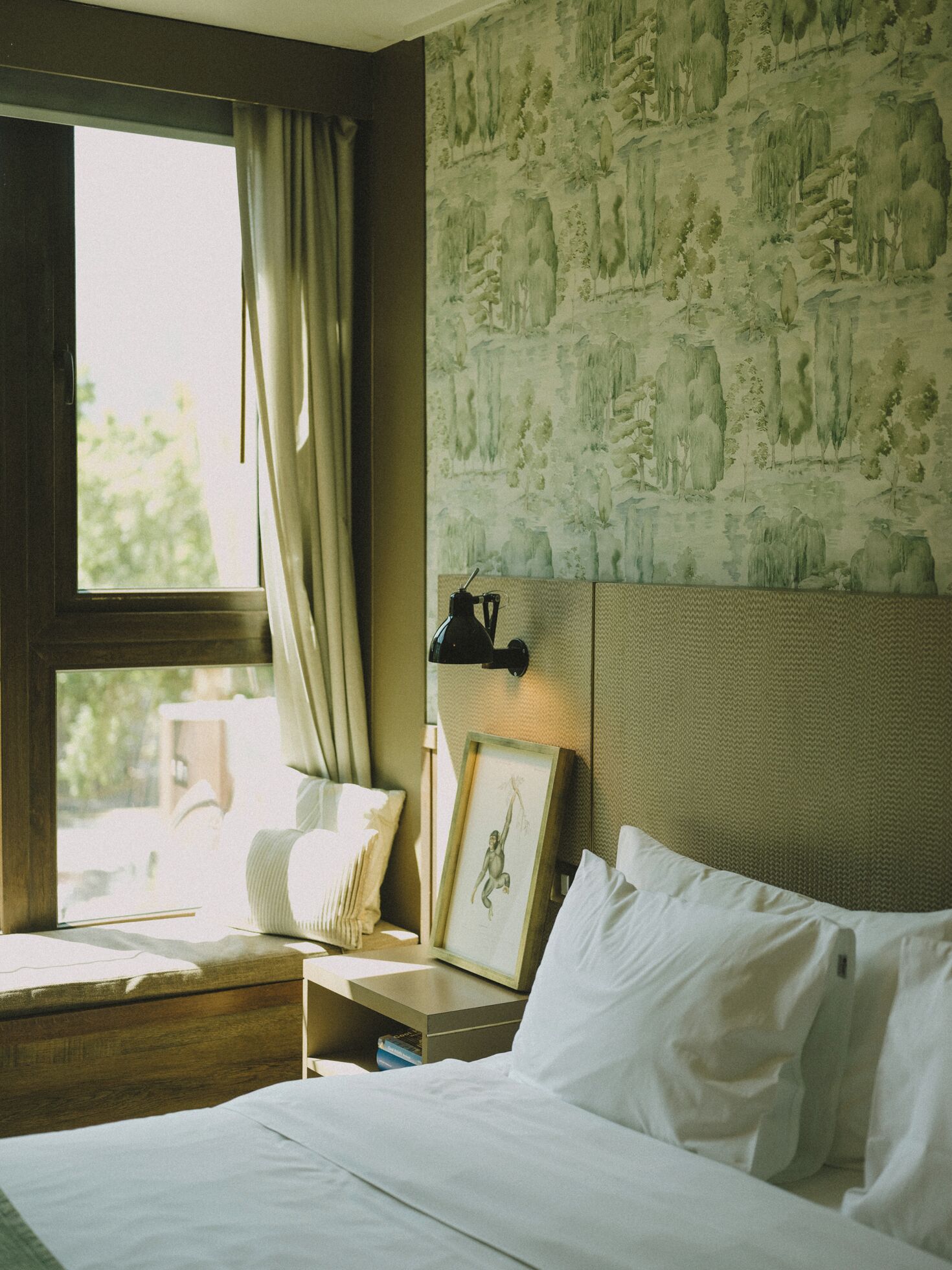 A hotel room with a bed, a nightstand, a lamp, a window with curtains, and a wall with a nature-themed mural.