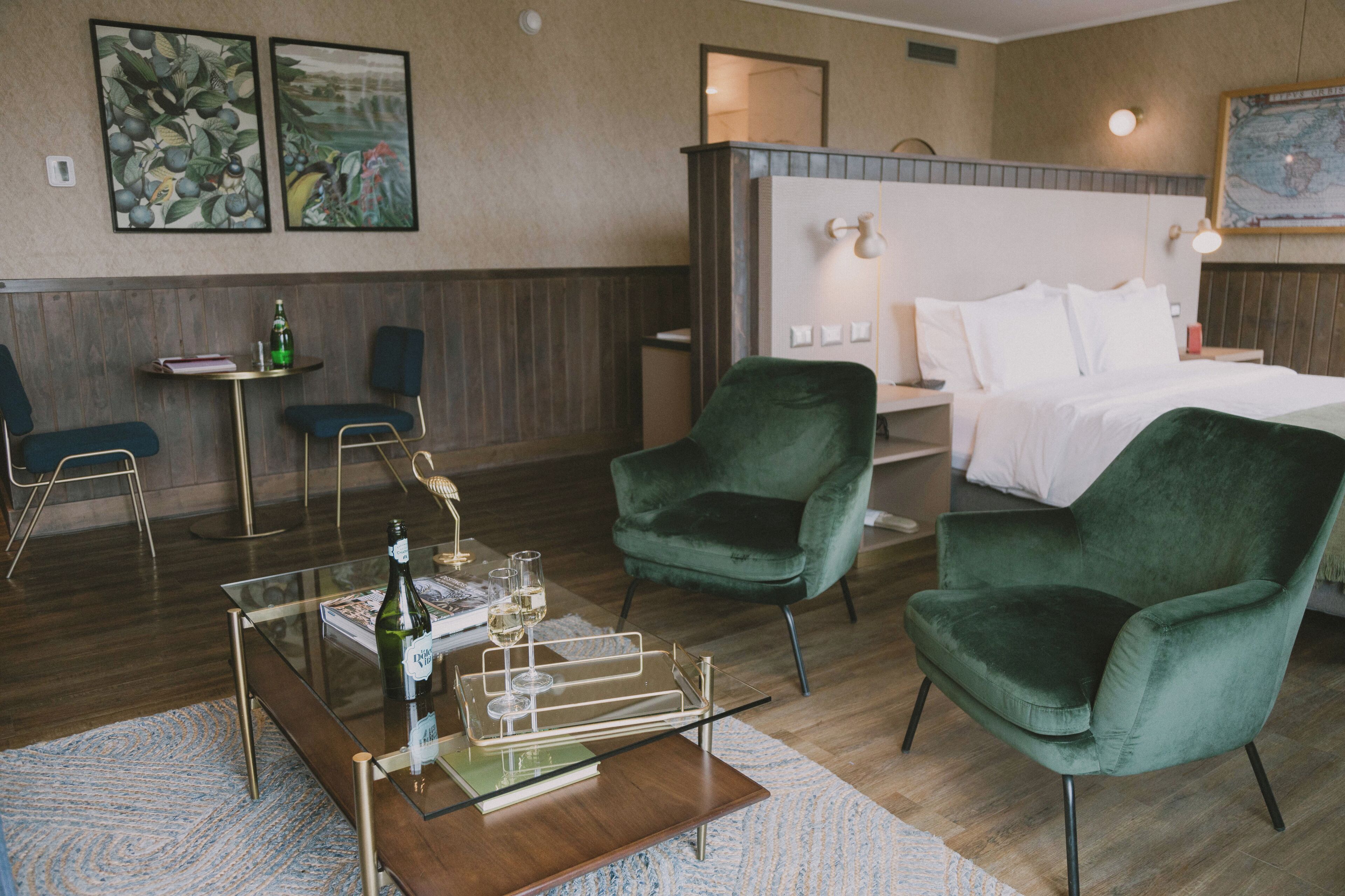 Una habitación de hotel con una cama, dos sillones verdes, una mesa de centro de vidrio con una botella y vasos, una mesita con un libro y una botella, y dos cuadros en la pared.