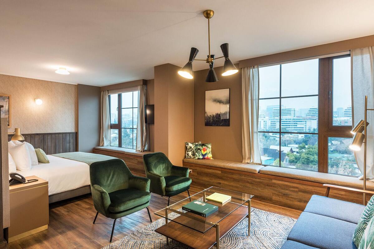 Habitación de hotel con una cama, dos sillones verdes, una mesa de centro de vidrio, un sofá, un escritorio con teléfono y ventanas amplias con vistas a la ciudad.