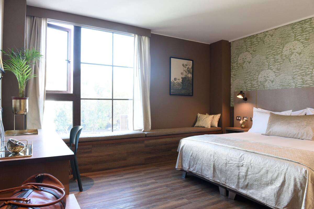 Habitación de hotel con una cama grande, un escritorio con silla, una ventana con cortinas, una planta y una lámpara de pared.