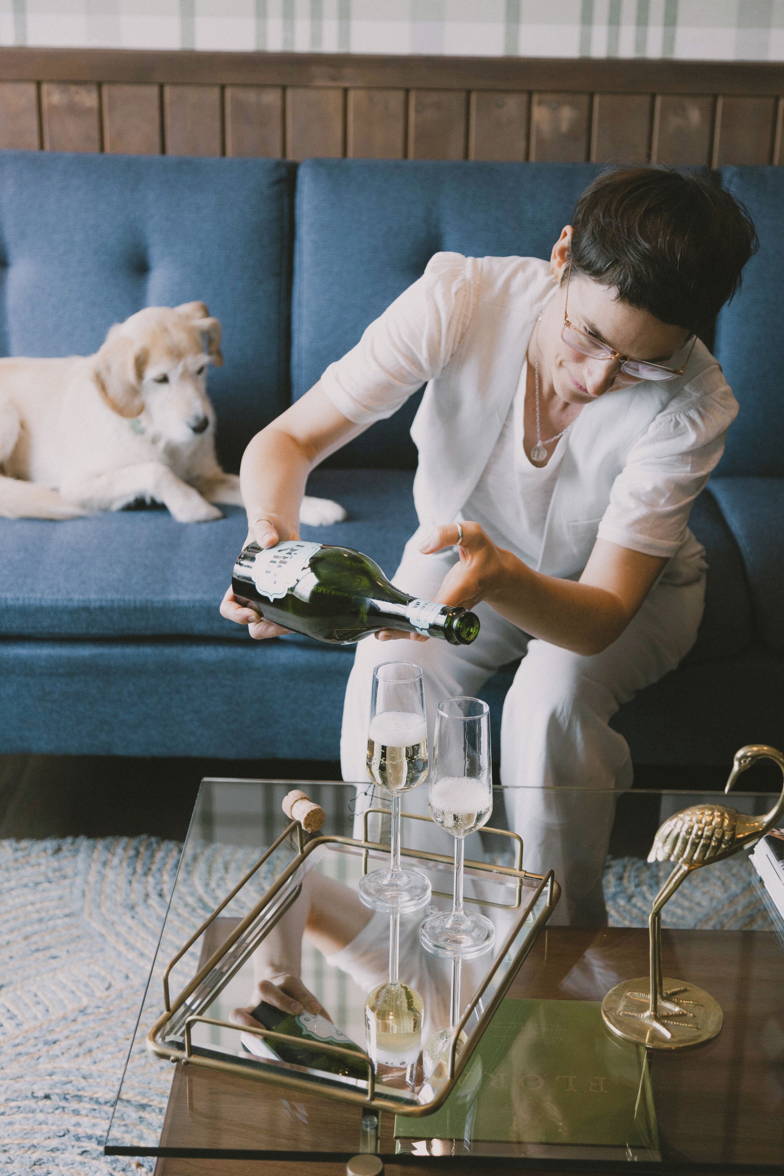 Eine Person gießt Champagner in Gläser auf einem Tablett, im Hintergrund liegt ein Hund auf einem blauen Sofa.