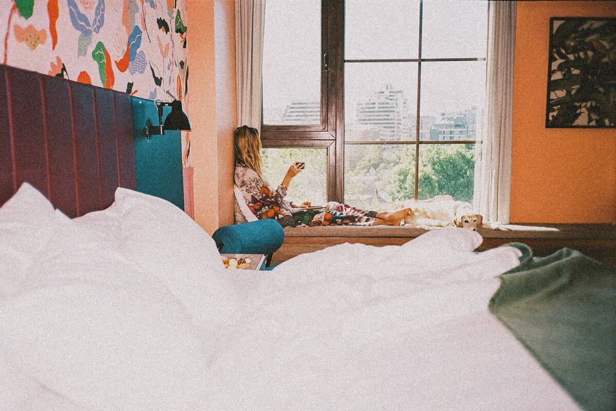 Una habitación con una cama, un ventanal con vistas a edificios y una pared de colores.