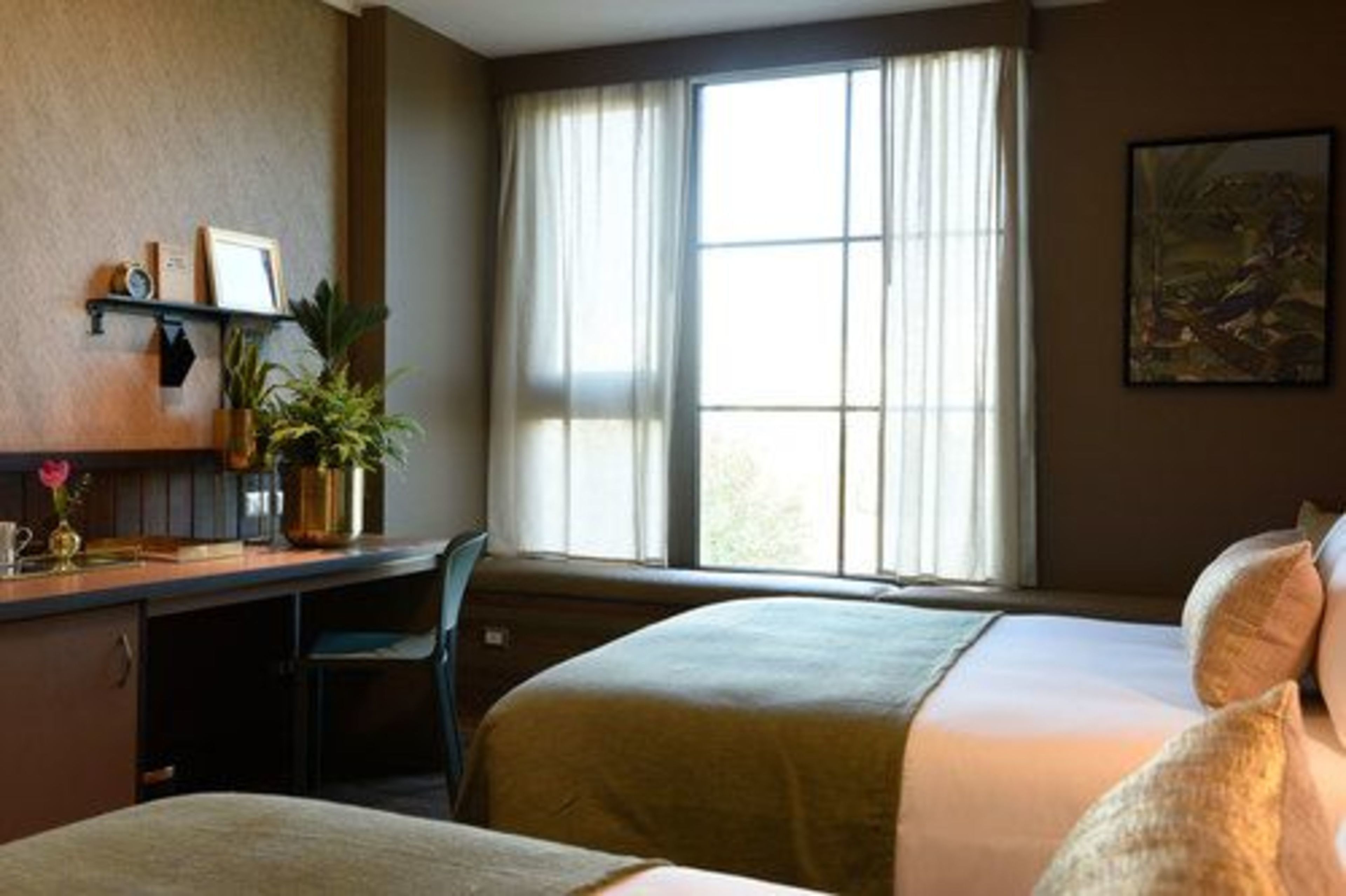 Een hotelkamer met twee bedden, een bureau met stoel, een raam met gordijnen en een plank met spullen.