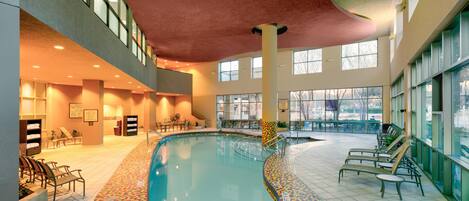 An indoor swimming pool with a colorful mosaic floor, surrounded by lounge chairs and a large window offering a view of the outdoors.