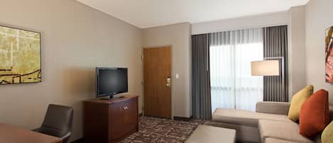 A hotel room with a flat-screen TV, a wooden cabinet, a sofa, a small table, and a large window with curtains.