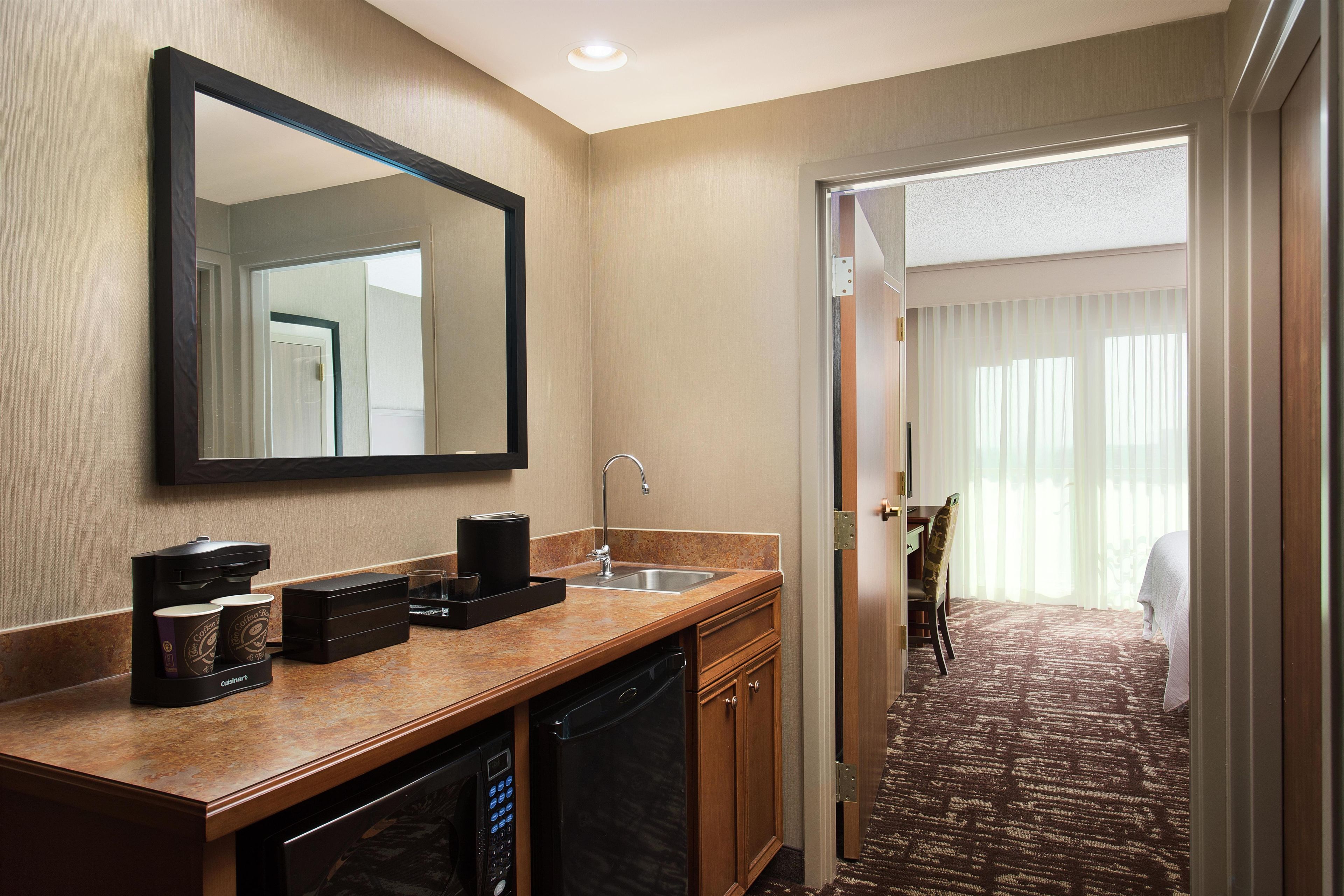 A hotel bathroom with a sink, mirror, coffee maker, and a view of a bedroom with a bed and curtains.
