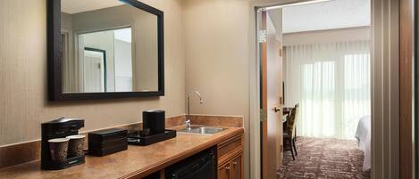 A hotel bathroom with a sink, mirror, coffee maker, and a view of a bedroom with a bed and curtains.