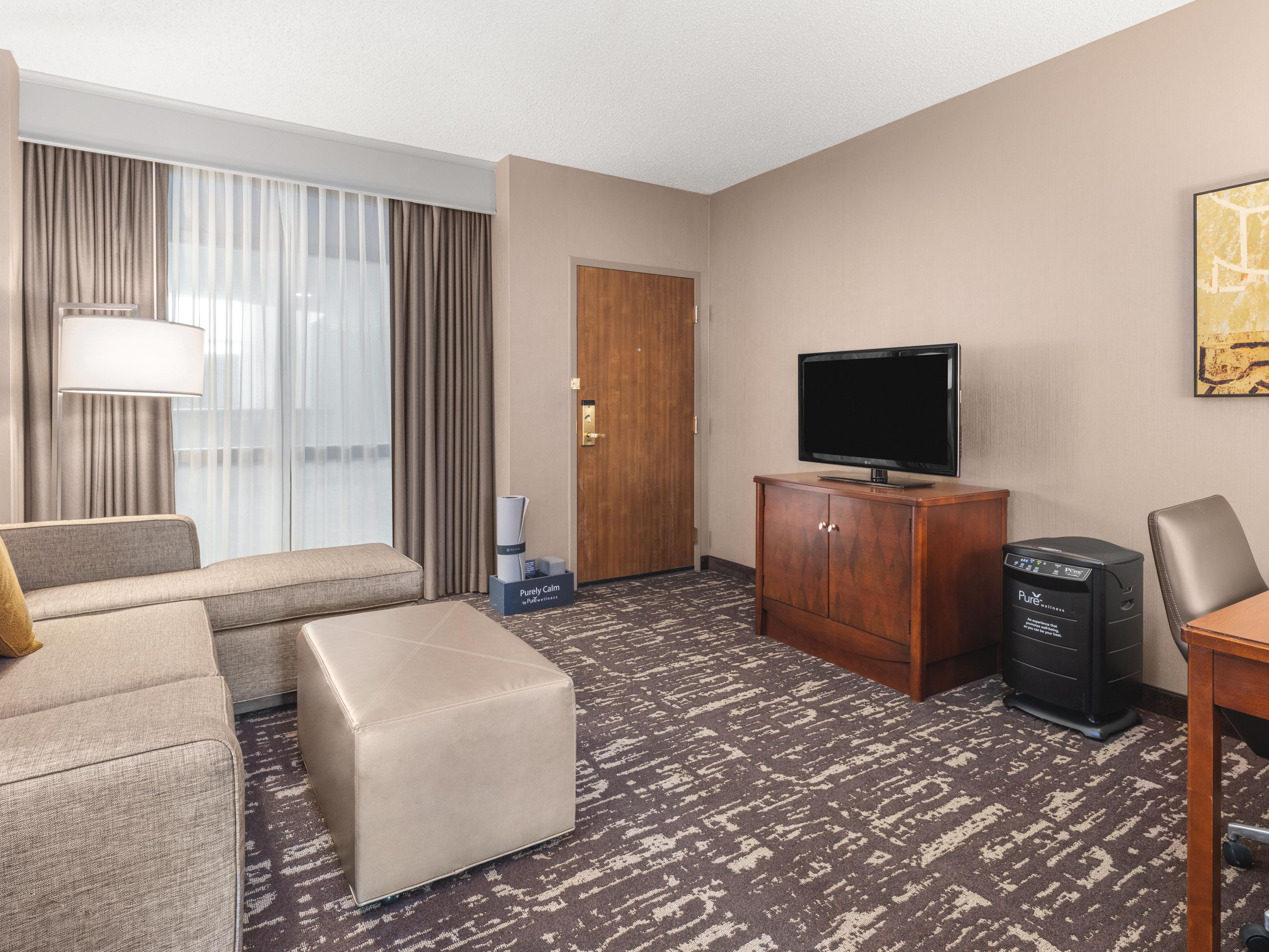 A hotel room with a sofa, ottoman, television, cabinet, and a desk with a computer.
