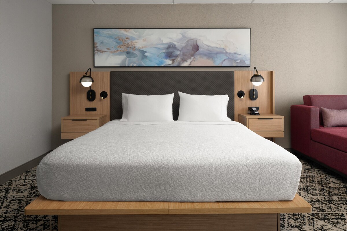 Quarto de hotel com cama grande, duas mesas de cabeceira com luminárias, uma poltrona vermelha e um quadro abstrato pendurado na parede.