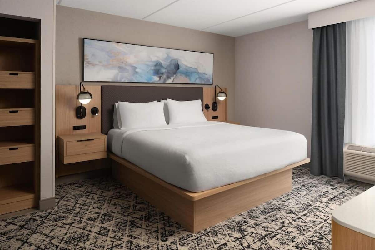 Habitación de hotel con una cama grande, cabecera de madera, mesitas de noche con lámparas, un cuadro abstracto colgado en la pared y una alfombra estampada.