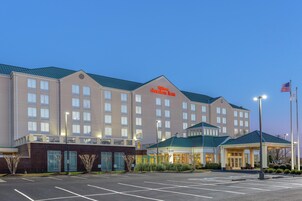 Un hotel grande con un letrero visible que dice 'Hilton Garden Inn', un estacionamiento y un asta con la bandera estadounidense.