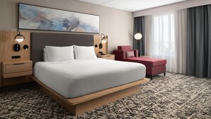 Een moderne hotelkamer met een groot bed, een rode fauteuil, een bureau en een groot raam met gordijnen.