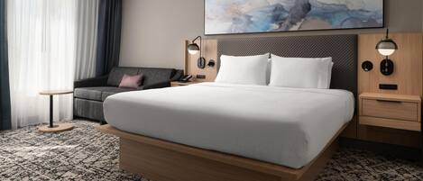 A hotel room with a large bed, a bedside table, a small desk, a chair, a sofa, and a painting on the wall.