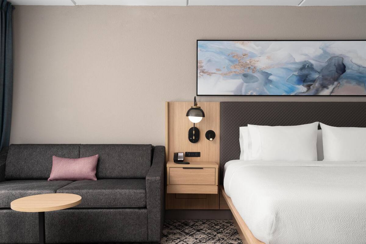 Habitación de hotel con sofá, una mesita, una mesita de noche con lámpara, cama y un cuadro colgado en la pared.