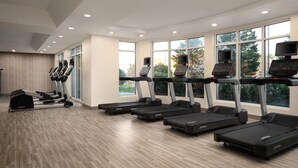 Een sportschool met loopbanden, elliptische trainers en een roeimachine, grote ramen en een houten vloer.