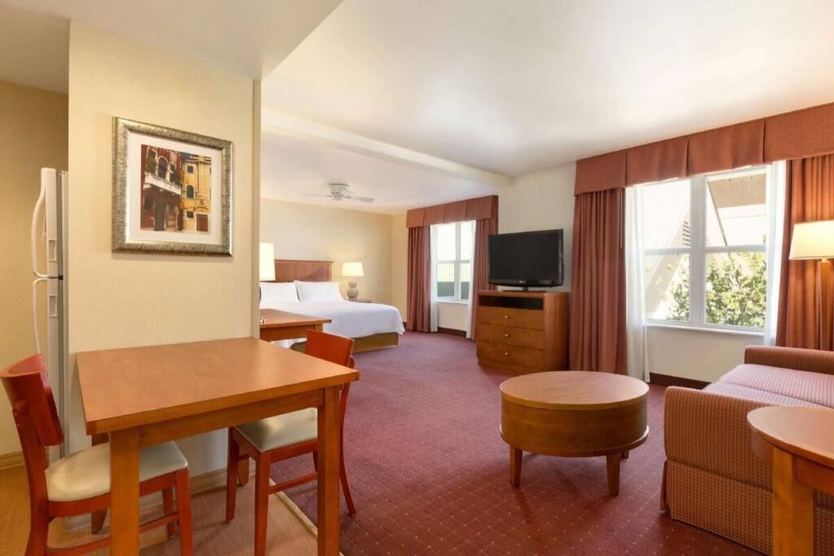 Ein Hotelzimmer mit Bett, Fernseher, Esstisch mit Stühlen, einer Couch und einem Fenster mit Vorhängen.