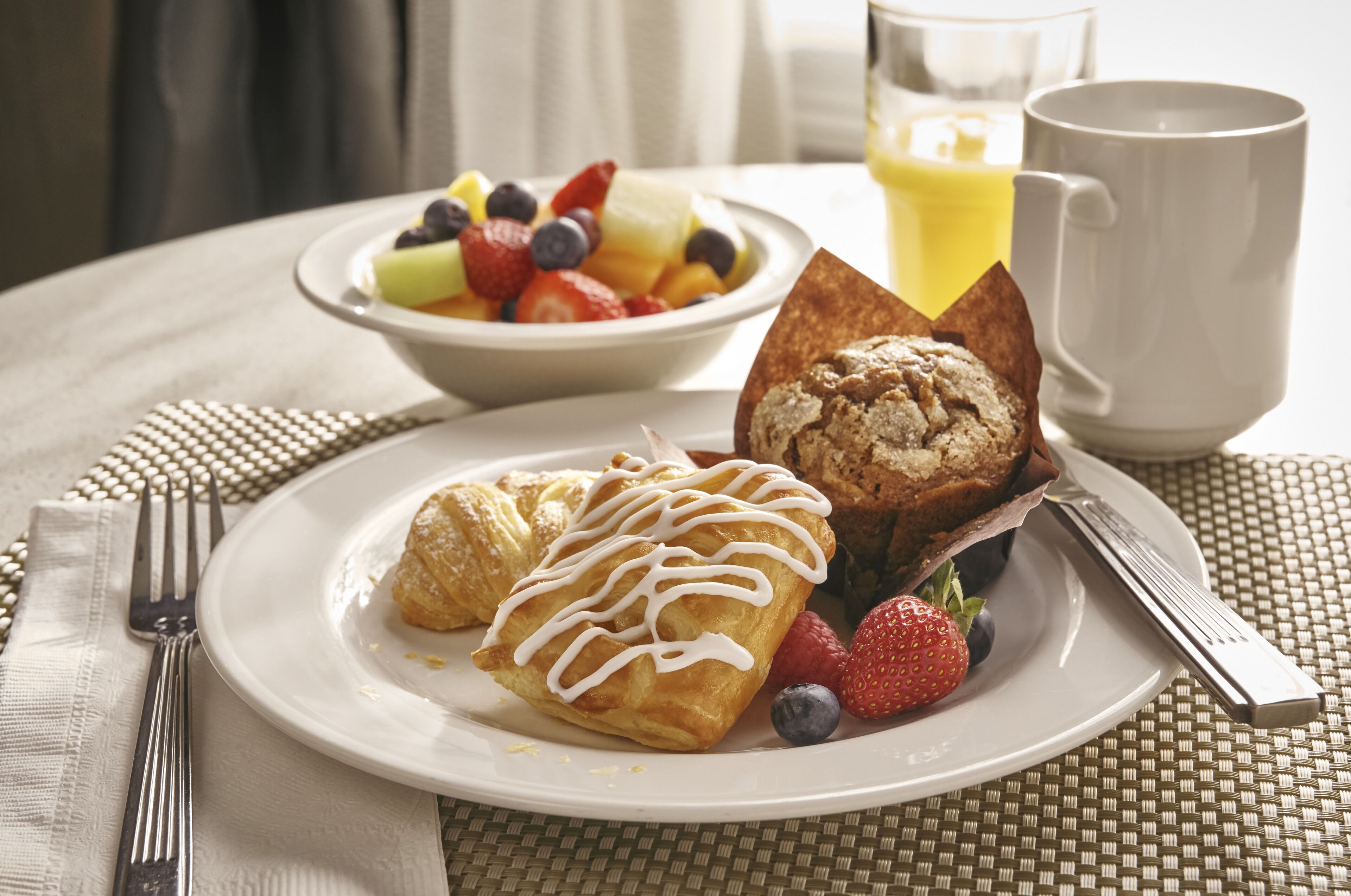 A breakfast setting with a plate of pastries, fresh berries, and a glass of orange juice on a woven tablecloth.