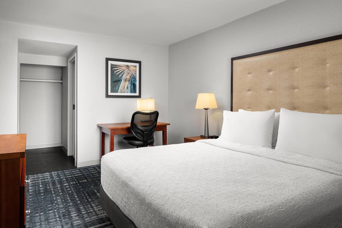 Ein Hotelzimmer mit einem großen Bett, einem Schreibtisch mit Stuhl, einer Lampe, einem gerahmten Bild an der Wand und einem Kleiderschrank.