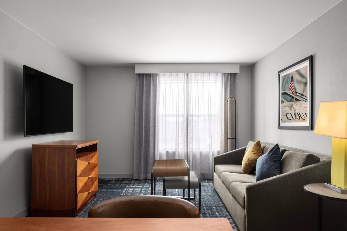 Ein modernes Hotelzimmer mit Flachbildfernseher, einem hölzernen Schminktisch, einer Couch mit Kissen, einem kleinen Tisch und einer Lampe.
