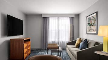 Una habitación de hotel moderna con televisión de pantalla plana, cómoda de madera, sofá con cojines, una mesita y una lámpara.