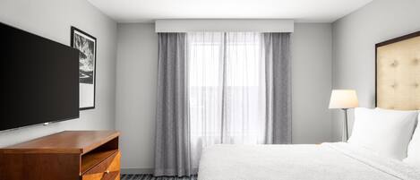 Habitación de hotel con cama, televisor de pantalla plana, ventilador de techo, ventana con cortinas y mesita de noche con lámpara.