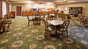 A hotel dining area with a carpeted floor, wooden tables and chairs, a counter with a coffee station, and a television. - Holiday Inn Express & Suites Thomasville by IHG (Thomasville)