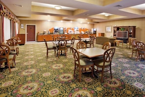A hotel dining area with a carpeted floor, wooden tables and chairs, a counter with a coffee station, and a television. - Holiday Inn Express & Suites Thomasville by IHG (Thomasville)