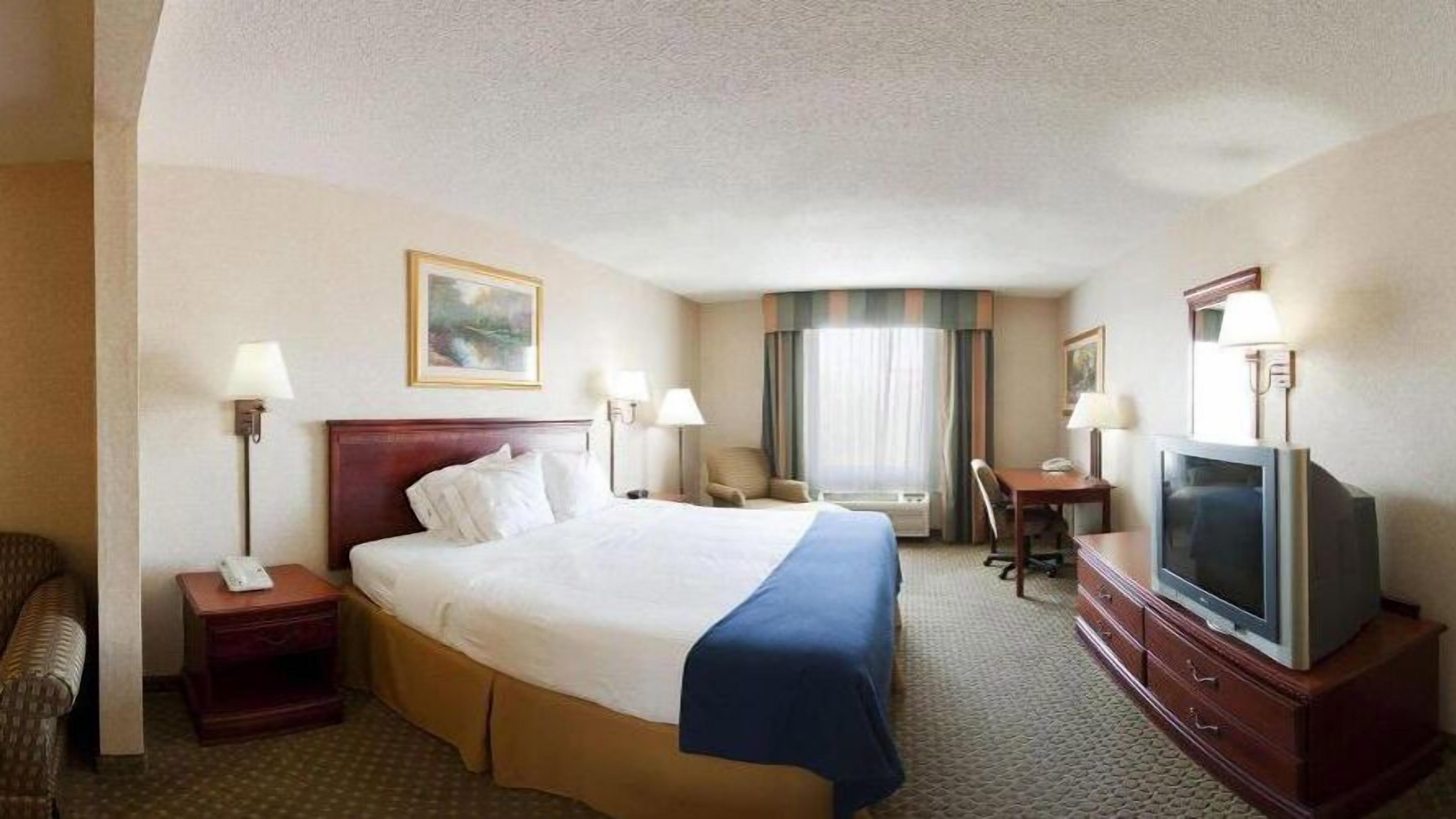 Ein Hotelzimmer mit einem großen Bett, einem Schreibtisch mit Stuhl, einem Fernseher und einem Fenster mit Vorhängen.