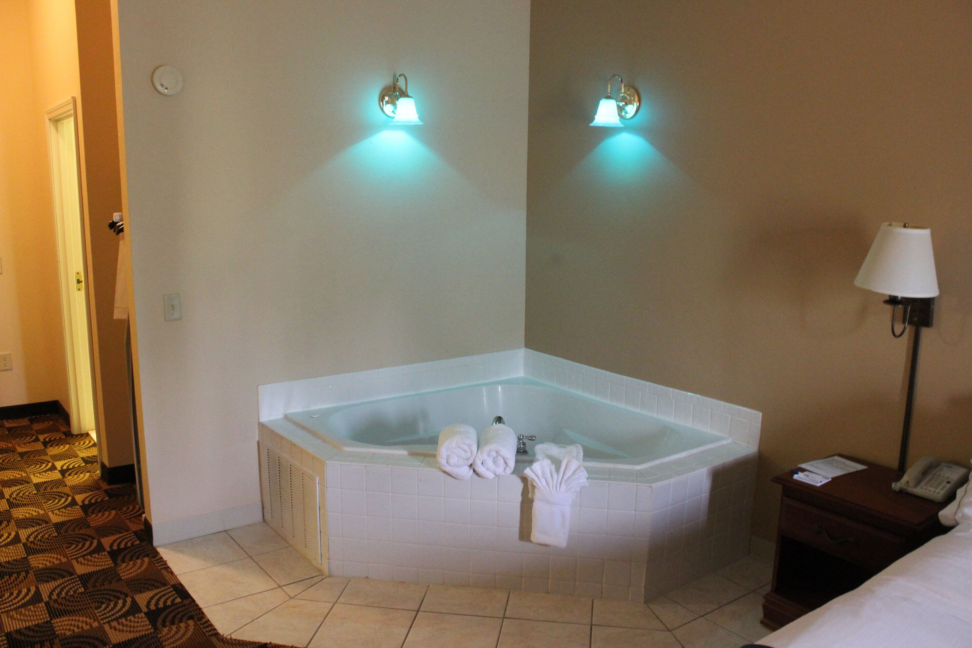 Ein Hotelzimmer mit Eckbadewanne, einem Nachttisch mit Lampe und einer Tür zu einem anderen Raum.