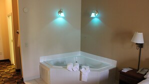 A hotel room with a corner bathtub, two wall-mounted lights, a bedside table with a lamp, and a telephone.