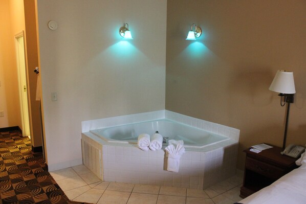 Habitación de hotel con bañera de hidromasaje en esquina, dos luces fijadas a la pared, una mesita de noche con lámpara y teléfono.