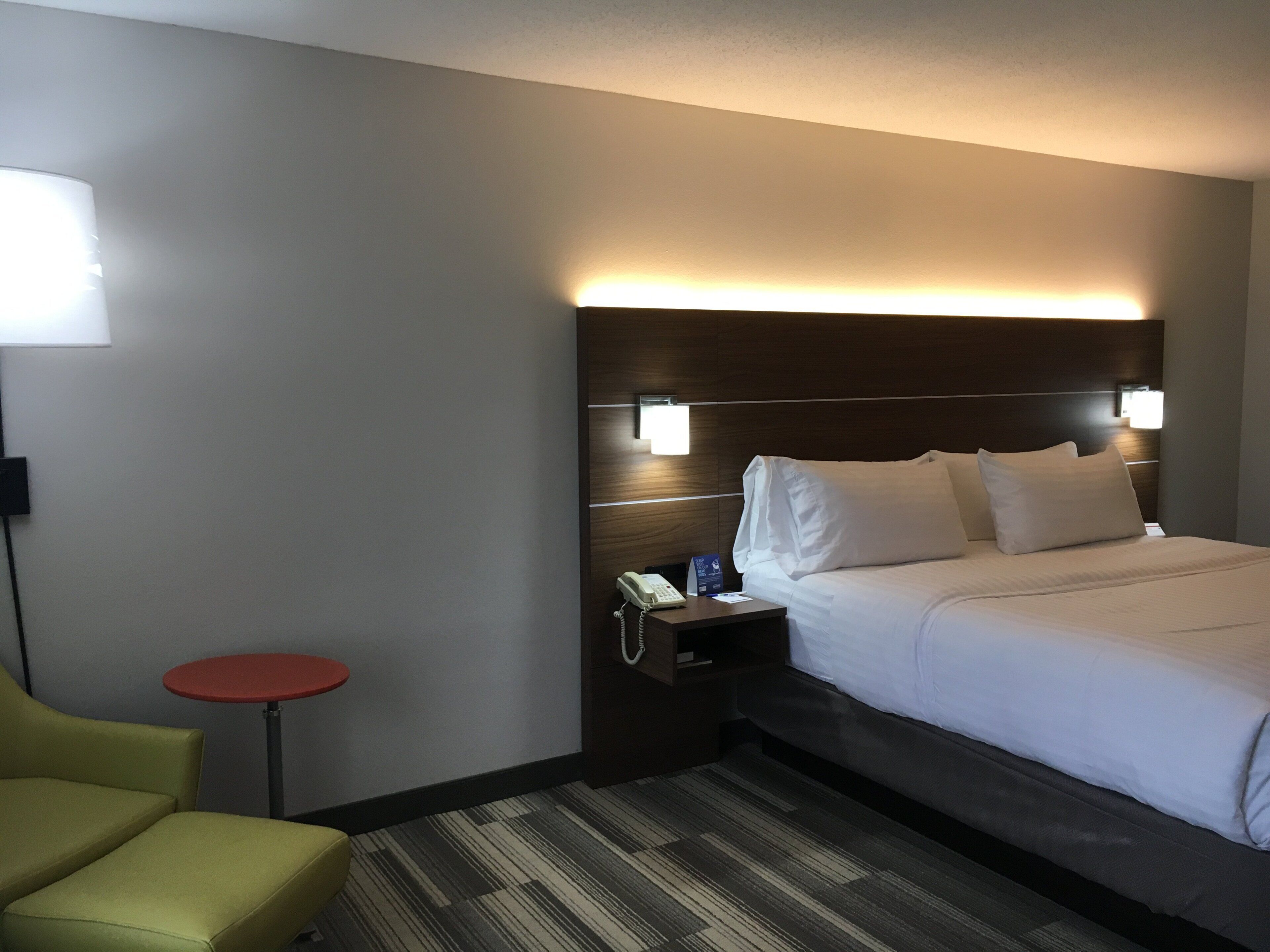 A hotel room with a bed, bedside table, lamp, and a small red table.