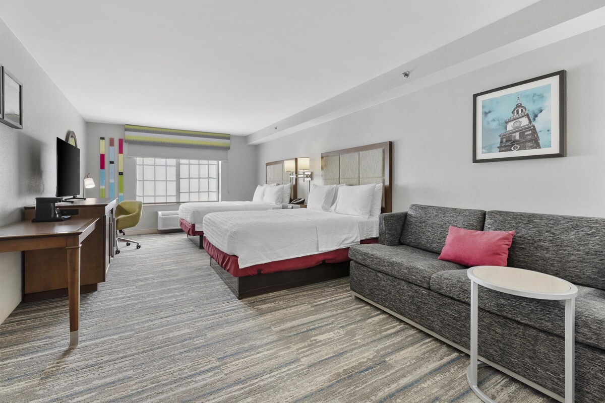 Habitación de hotel con escritorio, dos camas, sofá, una mesita y un cuadro en la pared.