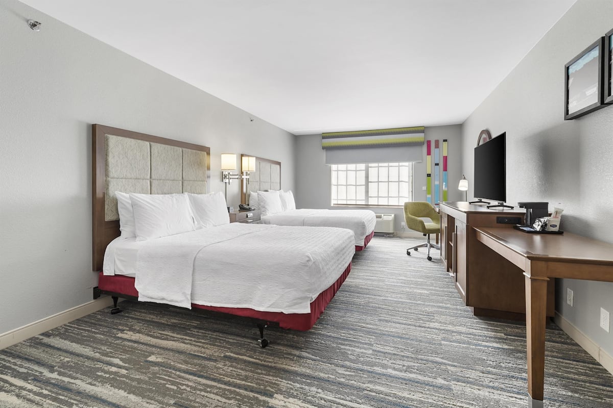 A hotel room with two beds, a desk, a TV, and a window.