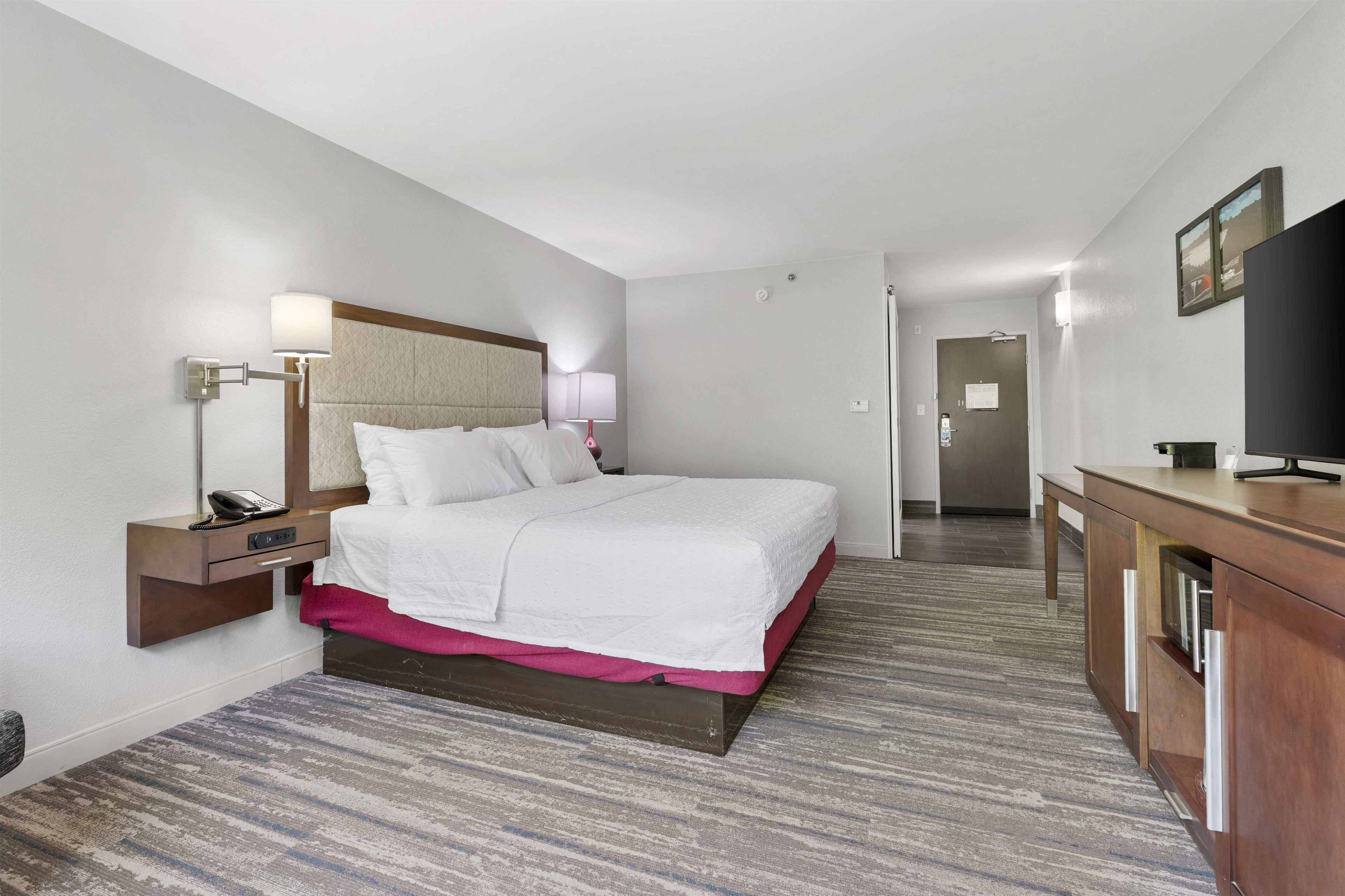 Una habitación de hotel con cama, mesita de noche, lámpara y televisor sobre un mueble de madera.