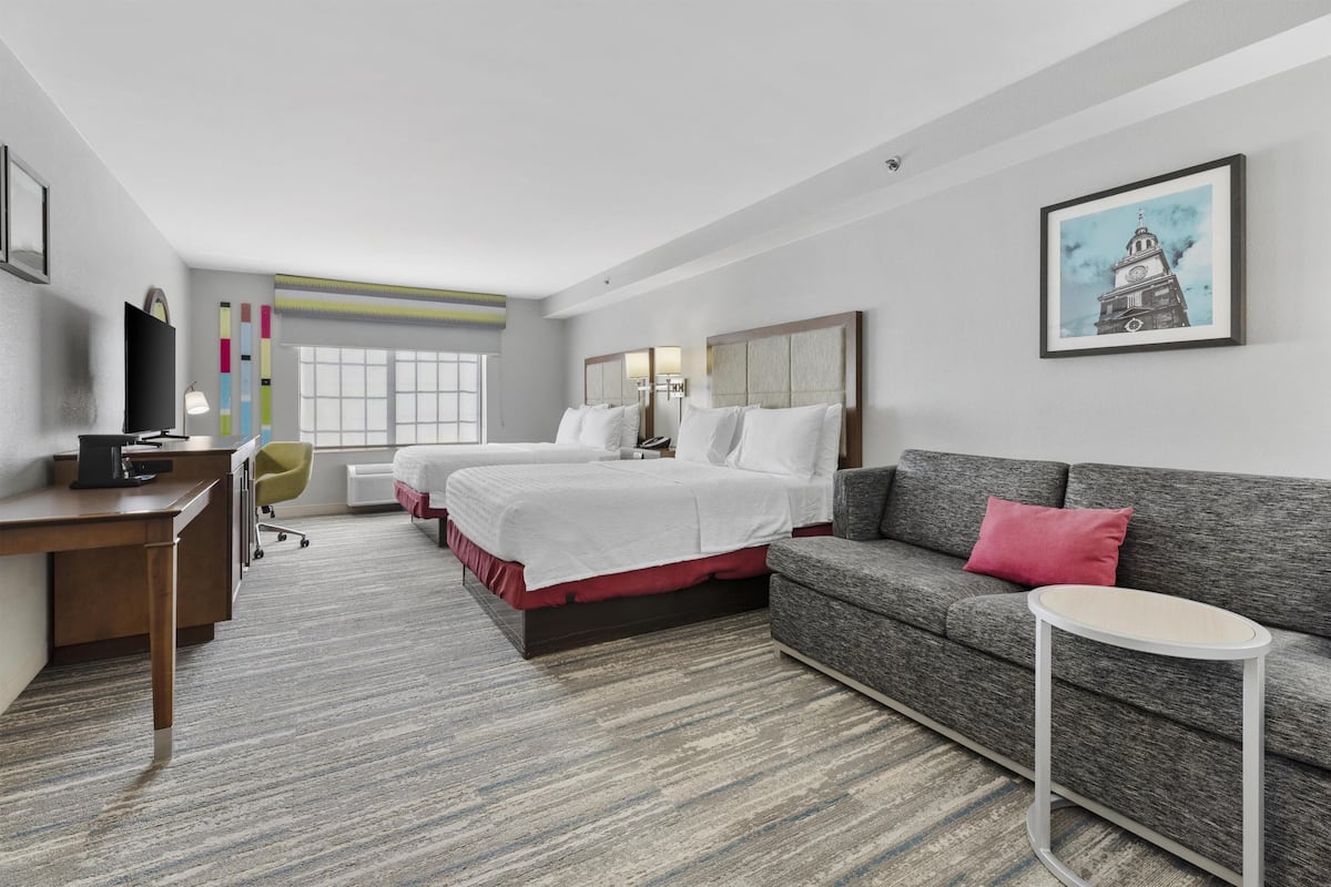 A hotel room with a desk, two beds, a sofa, a small table, and a painting on the wall.
