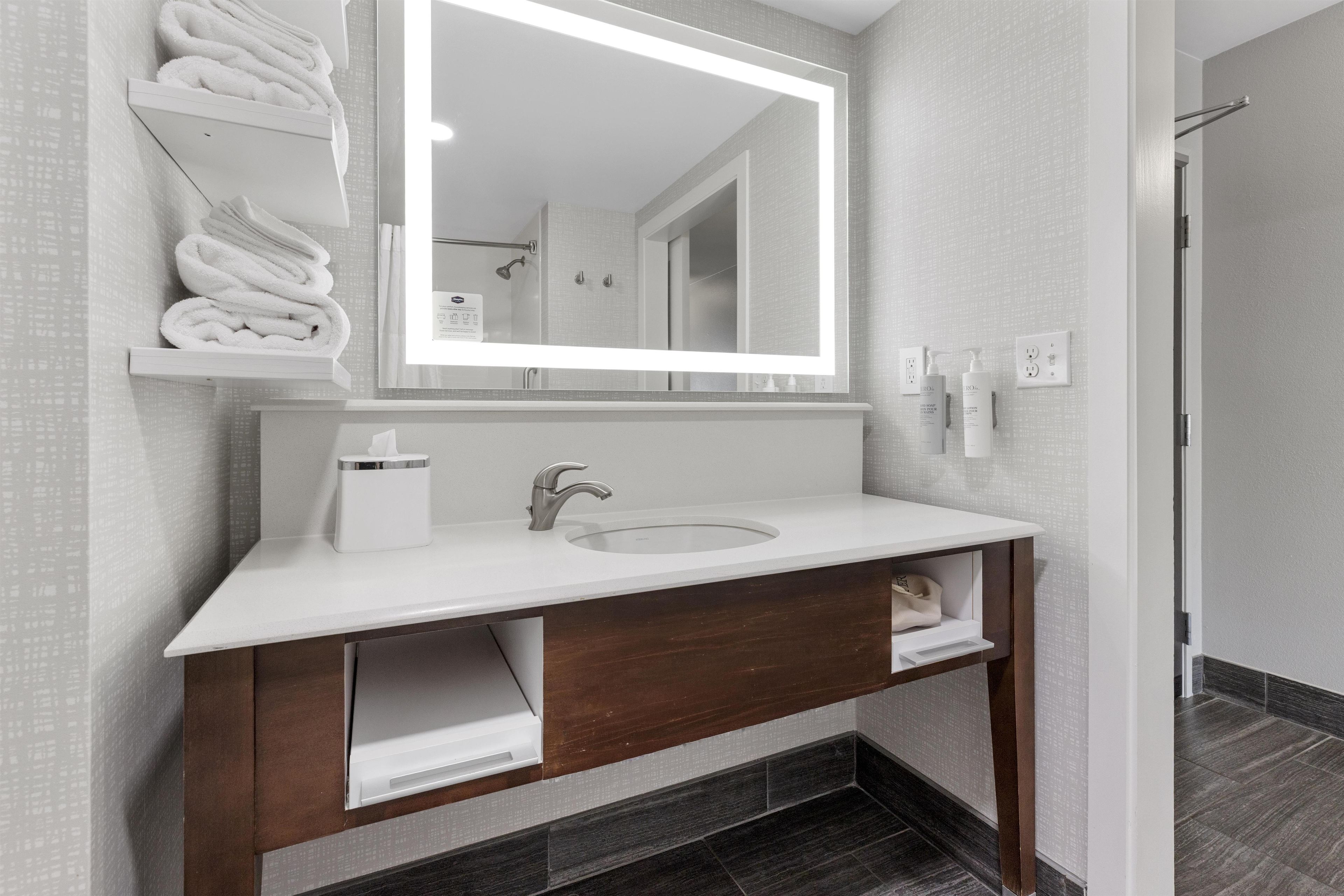 Un baño moderno con un lavamanos blanco, tocador de madera oscura, un espejo grande y toallas dobladas ordenadamente en un estante.