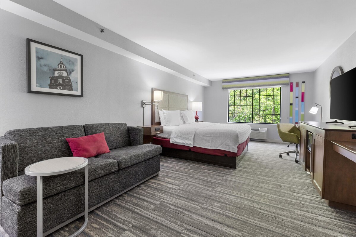 Habitación de hotel con cama, sofá, escritorio, silla, una mesita, televisor y ventana con vista a follaje.