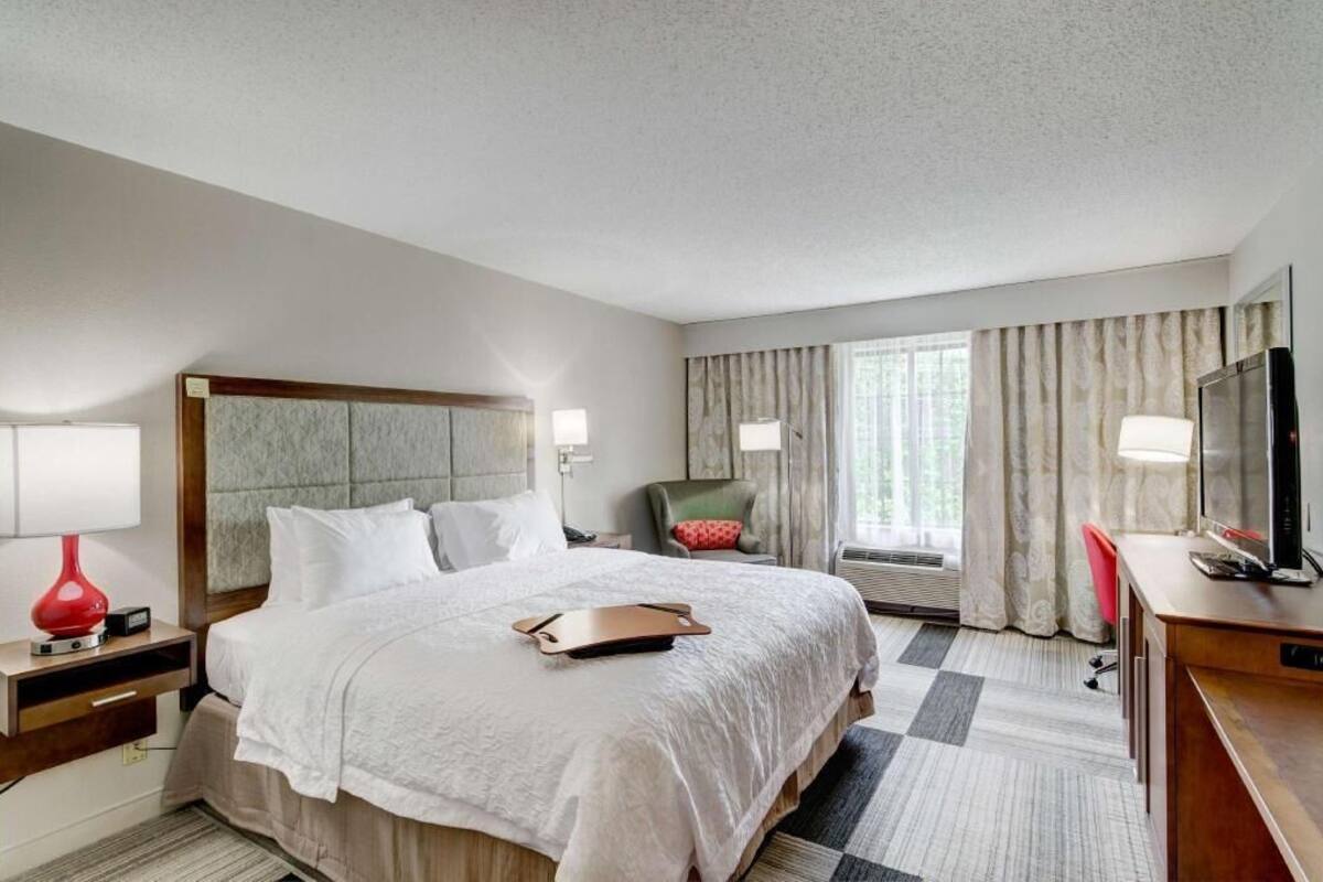 A hotel room with a bed, a desk with a television, a chair, and a window with curtains.