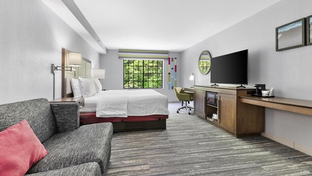 Une chambre d’hôtel équipée d’un lit, d’un bureau, d’une télévision et d’une fenêtre donnant sur un paysage verdoyant.