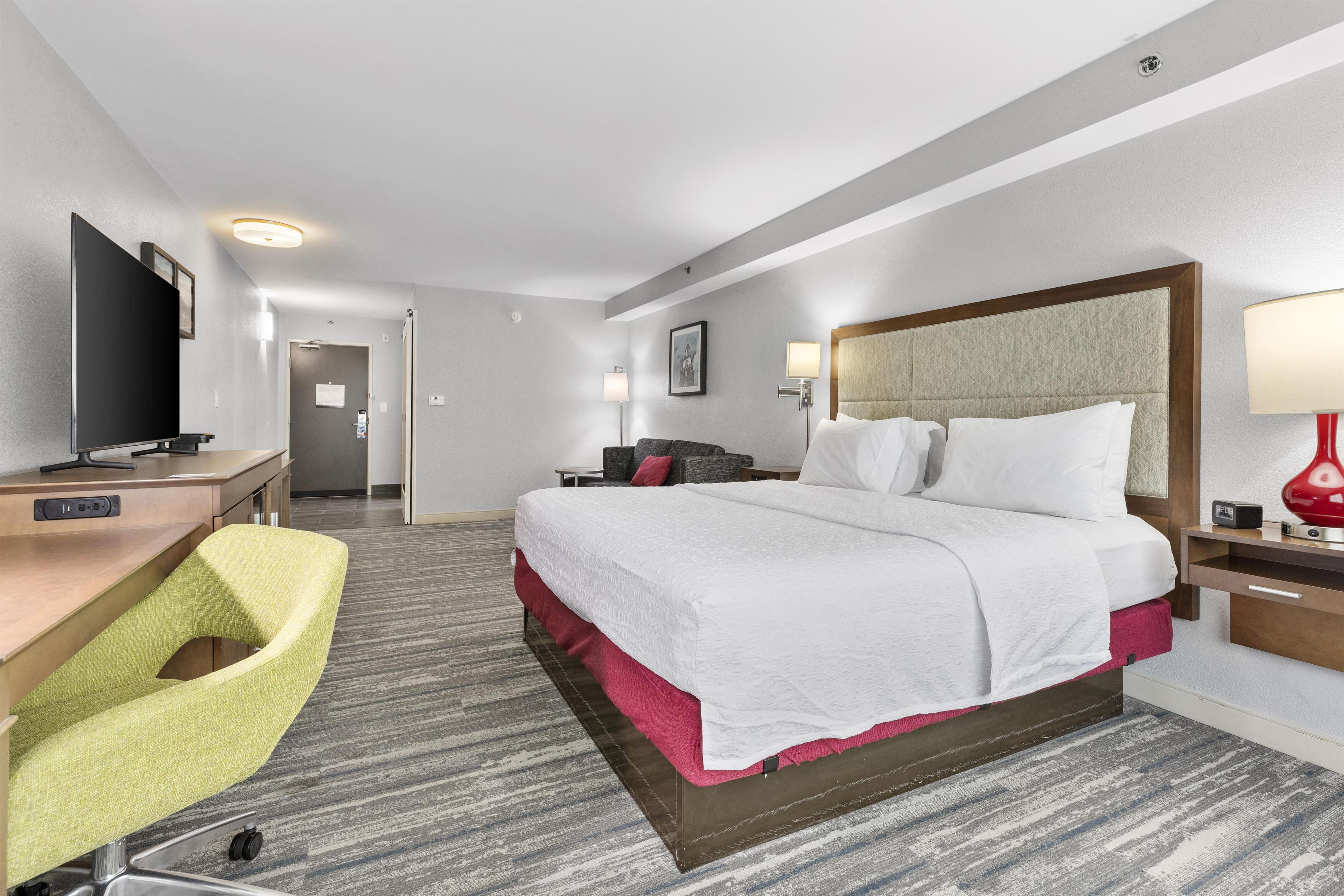 A hotel room with a bed, desk, chair, TV, bedside lamps, and a door leading to another room.