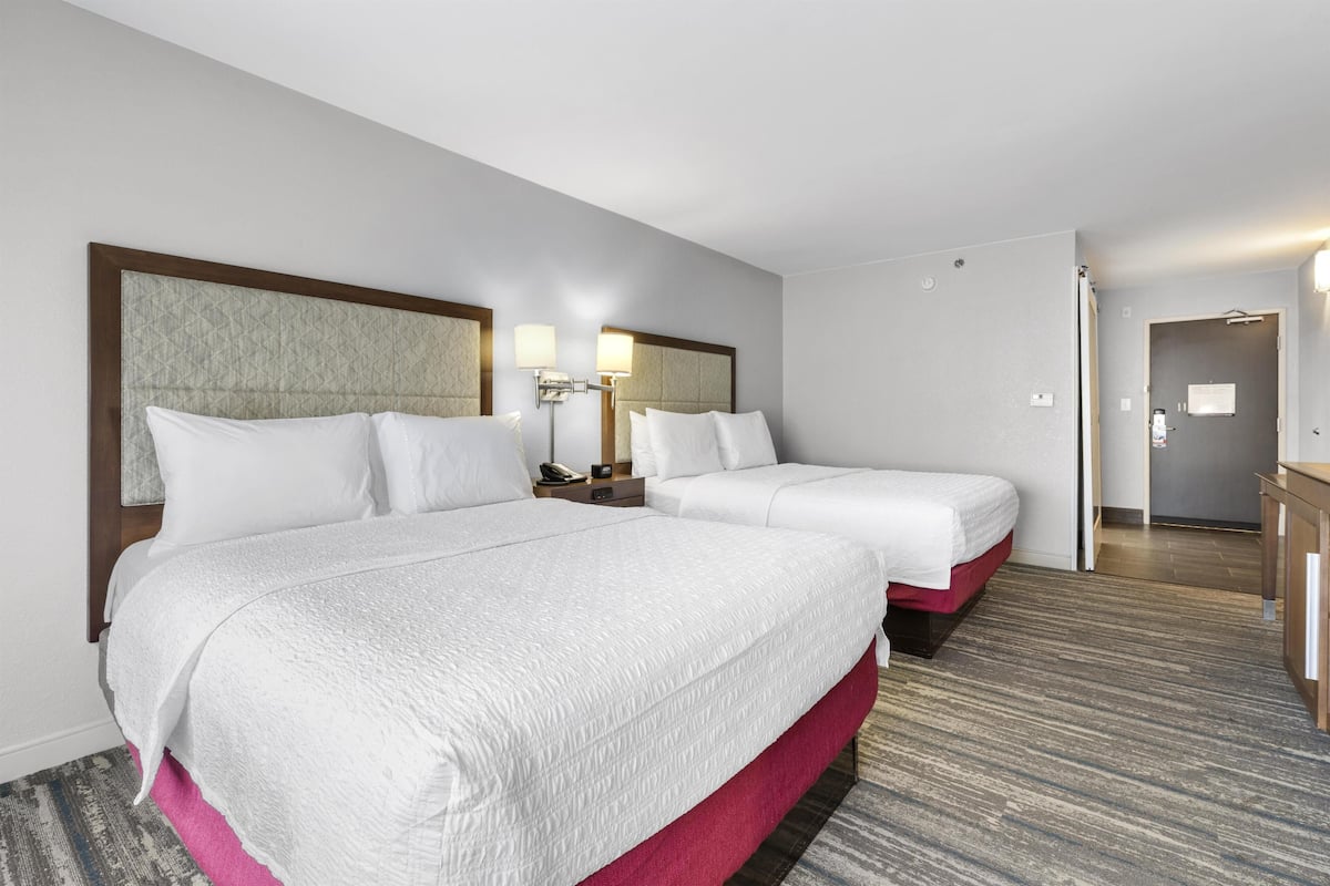 A hotel room with two beds, a nightstand, a lamp, and a door leading to a bathroom.