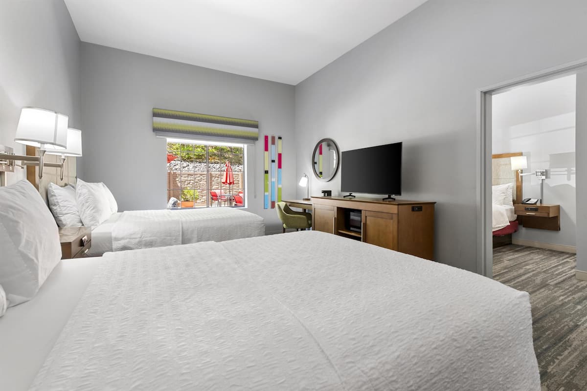 A hotel room with two beds, a desk, a TV, and a window with a view of the outside.