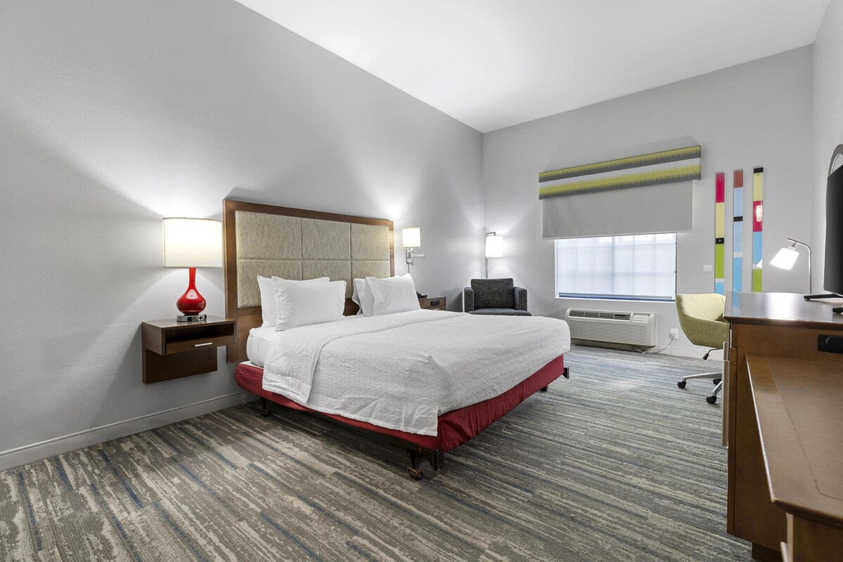 Habitación de hotel con una cama grande, mesitas de noche, un escritorio, una silla y una ventana con persianas.