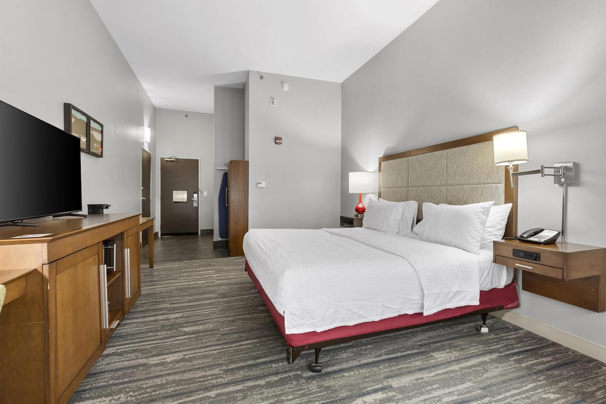 A hotel room with a bed, bedside table, lamp, TV, and a desk with drawers.