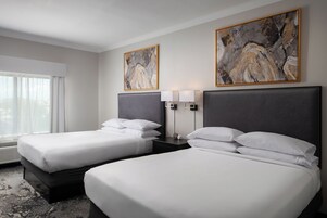 Quarto de hotel com duas camas, uma janela grande com cortinas leves e duas pinturas abstratas nas paredes.
