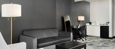 Une chambre d’hôtel moderne avec un canapé gris, un fauteuil blanc, une table basse noire, un bureau avec une chaise et un coin cuisine équipé d’un réfrigérateur et d’une machine à café.